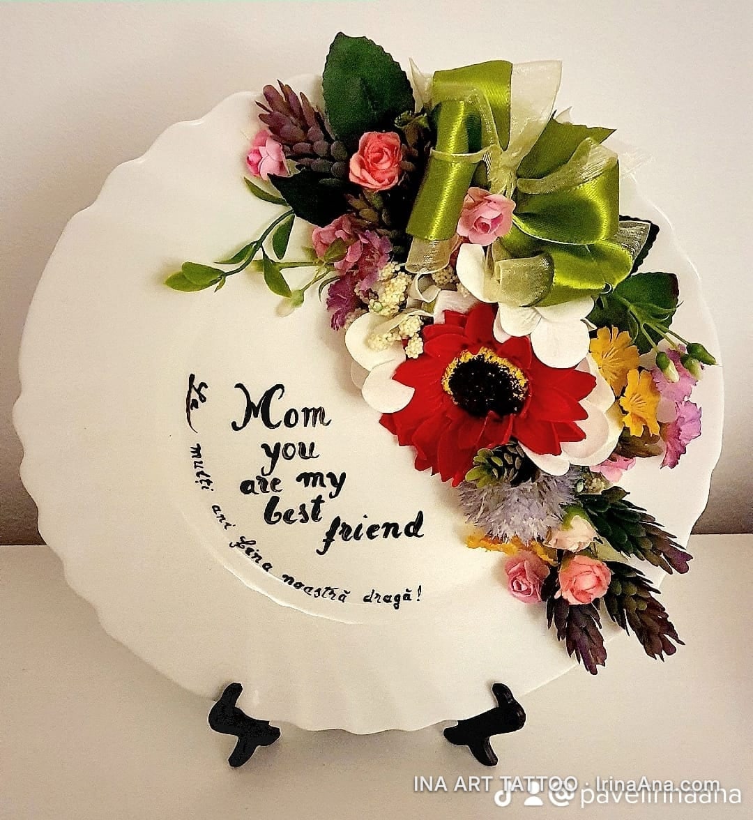 O farfurie decorativă albă, cu margine ondulată, decorată cu un aranjament floral și text caligrafic.