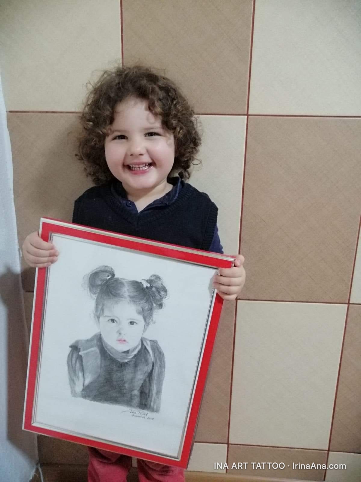 Un copil zâmbitor cu păr creț ține un portret în creion cu o fetiță cu cocuri, într-o ramă roșie.