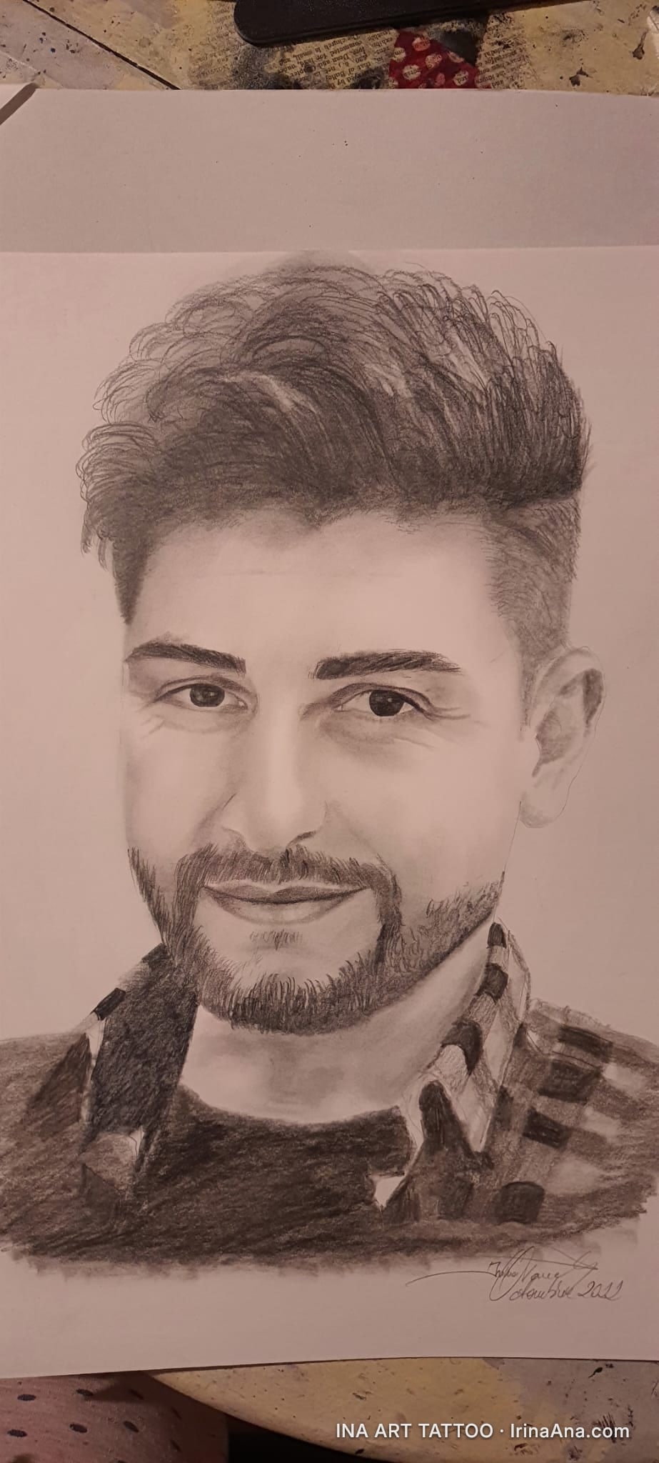 Portret în creion al unui bărbat tânăr, zâmbitor, cu barbă scurtă și păr tuns modern. Poartă o cămașă în carouri.